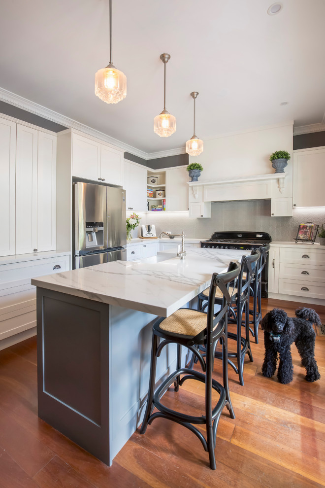 Mid-sized transitional l-shaped eat-in kitchen in Sydney with a farmhouse sink, shaker cabinets, white cabinets, quartz benchtops, grey splashback, metal splashback, black appliances, medium hardwood floors, with island, brown floor and multi-coloured benchtop.