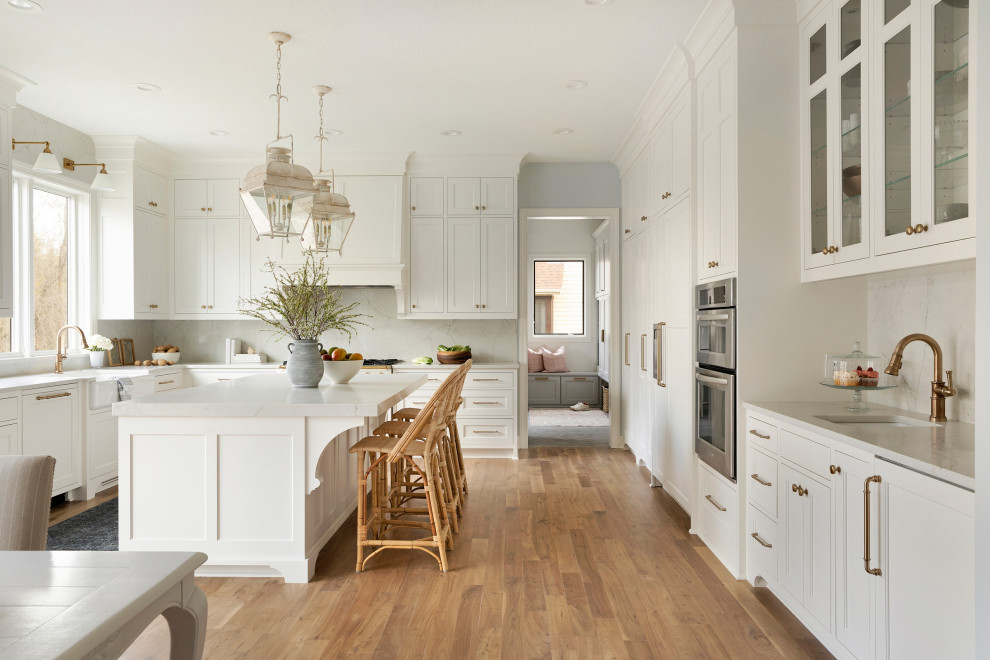 Photo of a large transitional l-shaped eat-in kitchen in Minneapolis with a farmhouse sink, beaded inset cabinets, white cabinets, quartzite benchtops, white splashback, engineered quartz splashback, panelled appliances, light hardwood floors, with island, brown floor and white benchtop.