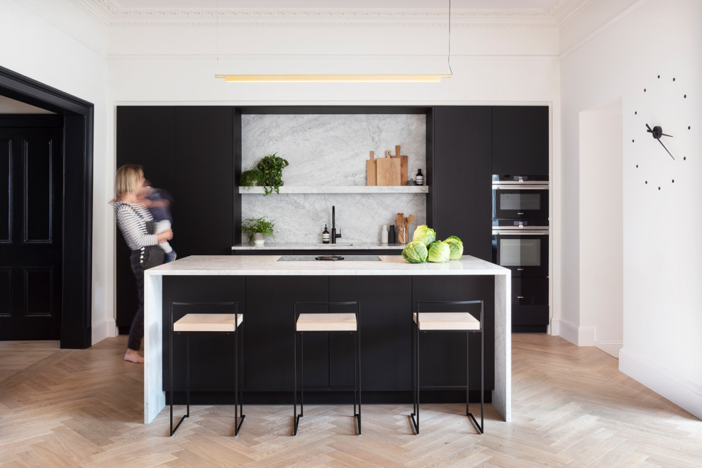 This is an example of a mid-sized scandinavian single-wall eat-in kitchen in Edinburgh with an undermount sink, flat-panel cabinets, black cabinets, marble benchtops, grey splashback, marble splashback, black appliances, light hardwood floors, with island, brown floor, grey benchtop and wallpaper.