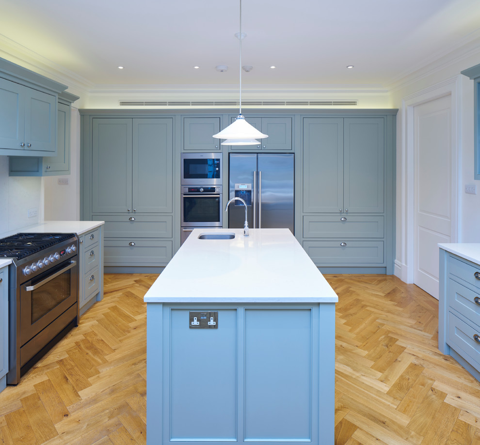 This is an example of a large traditional l-shaped open plan kitchen in London with a double-bowl sink, recessed-panel cabinets, grey cabinets, solid surface benchtops, white splashback, stainless steel appliances, medium hardwood floors and multiple islands.