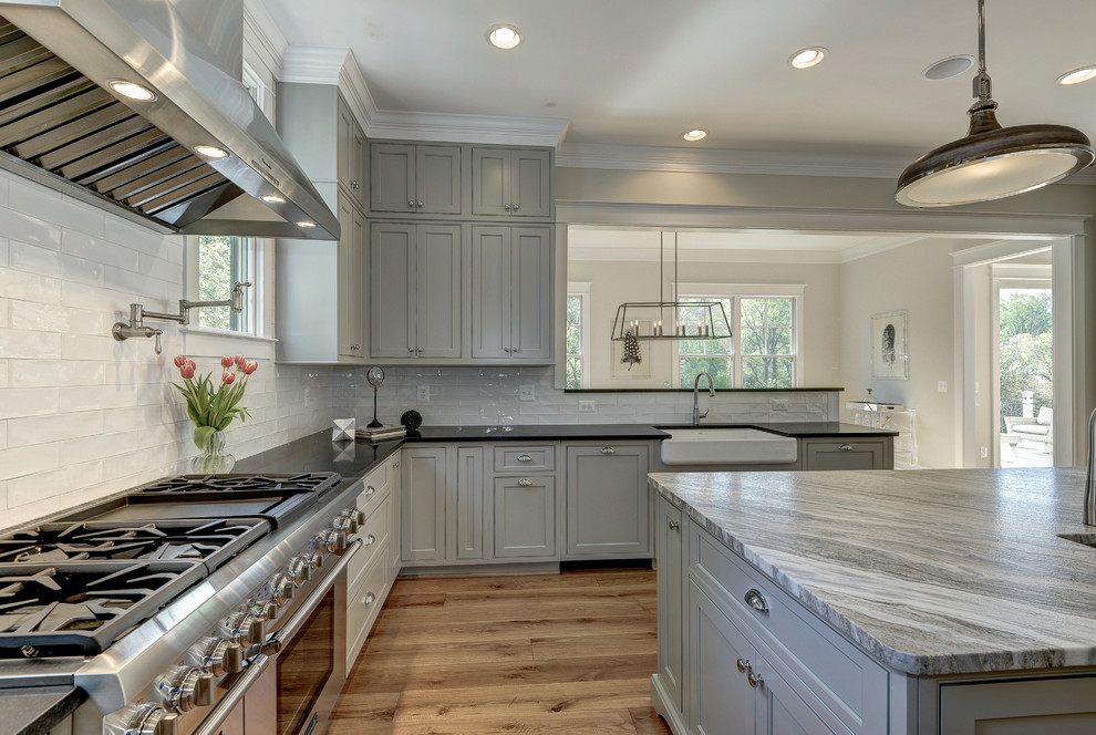 Design ideas for an expansive modern l-shaped kitchen in DC Metro with a farmhouse sink, raised-panel cabinets, grey cabinets, marble benchtops, white splashback, subway tile splashback, stainless steel appliances, light hardwood floors and with island.