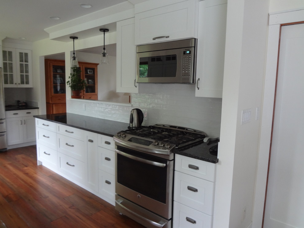 Example of a mid-sized transitional galley medium tone wood floor and brown floor kitchen design with an undermount sink, shaker cabinets, white cabinets, granite countertops, white backsplash, ceramic backsplash and stainless steel appliances