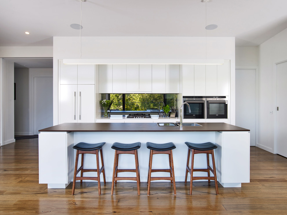 Inspiration for a large contemporary galley kitchen in Other with flat-panel cabinets, white cabinets, solid surface benchtops, metallic splashback, mirror splashback, black appliances, medium hardwood floors, with island, brown floor, brown benchtop and a drop-in sink.
