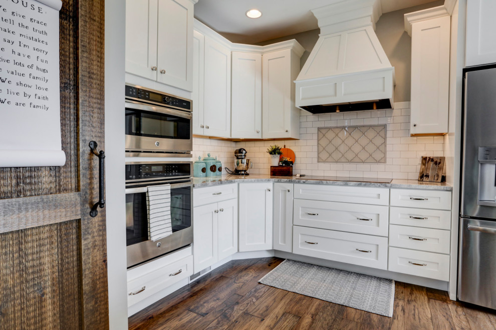 Country u-shaped kitchen with a farmhouse sink, white cabinets, quartzite benchtops, white splashback, subway tile splashback, stainless steel appliances, medium hardwood floors, with island, brown floor and grey benchtop.