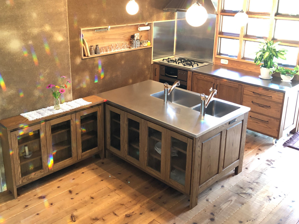 World-inspired galley kitchen/diner in Other with a double-bowl sink, beaded cabinets, medium wood cabinets, stainless steel worktops, wood splashback, light hardwood flooring, an island and brown floors.