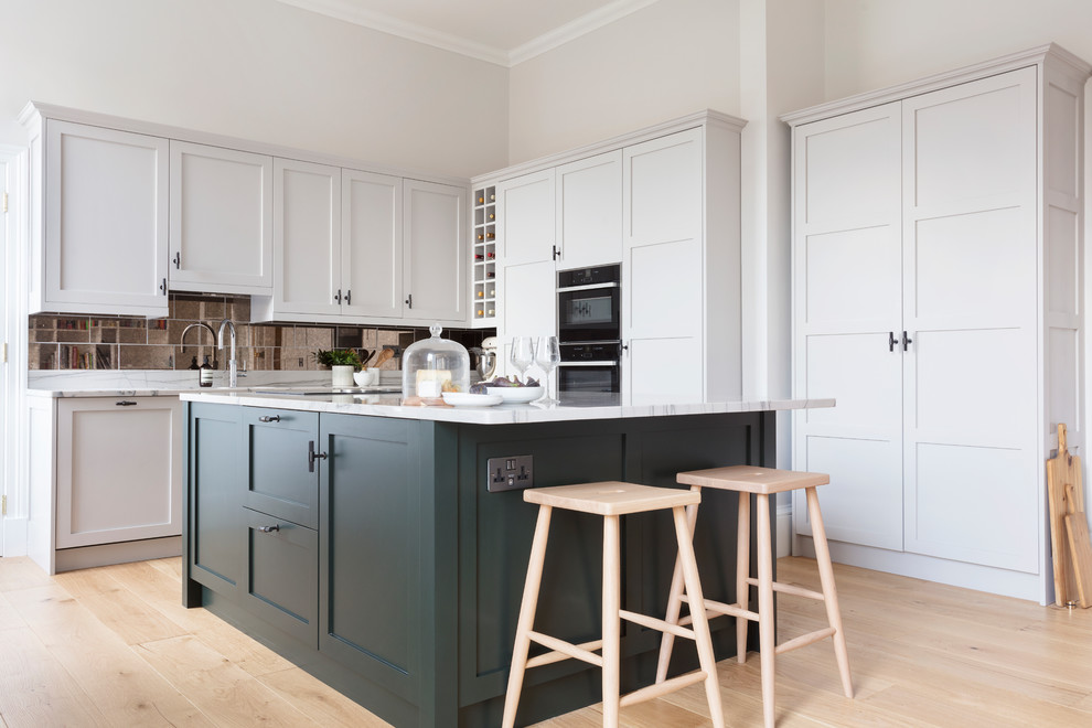 Design ideas for a mid-sized transitional l-shaped kitchen in Edinburgh with a drop-in sink, shaker cabinets, green cabinets, granite benchtops, metallic splashback, mirror splashback, black appliances, with island, white benchtop, light hardwood floors and beige floor.