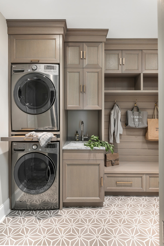 Design ideas for a mid-sized transitional single-wall utility room in Chicago with recessed-panel cabinets, grey cabinets, quartzite benchtops, grey splashback, stone slab splashback, grey benchtop, an undermount sink, beige walls, a stacked washer and dryer and beige floor.