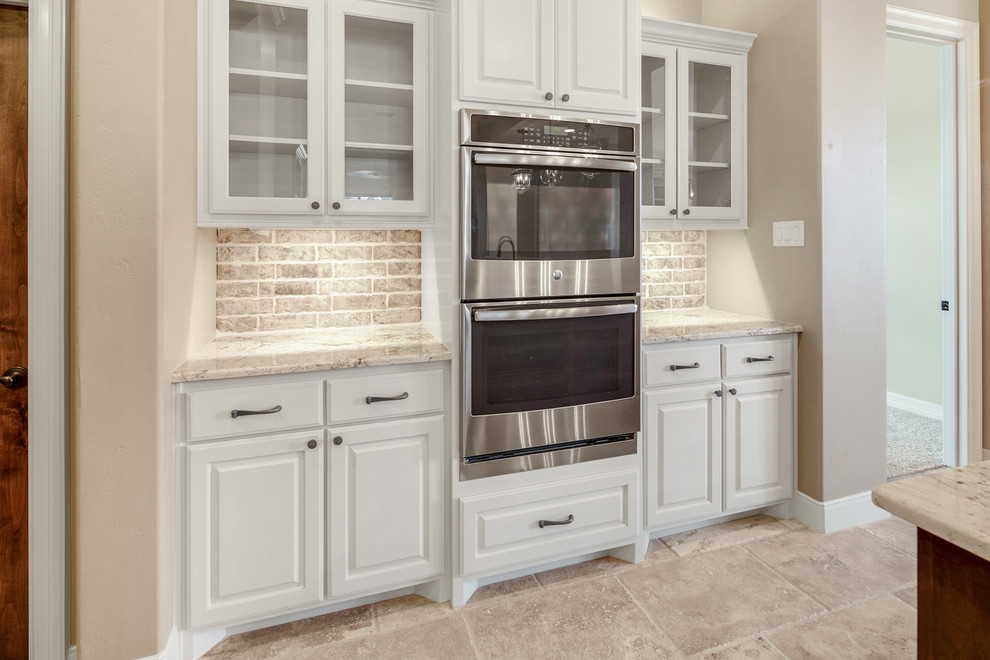 Inspiration for a large mediterranean kitchen pantry in Other with a farmhouse sink, raised-panel cabinets, beige cabinets, granite benchtops, beige splashback, brick splashback, stainless steel appliances, travertine floors, with island and beige floor.