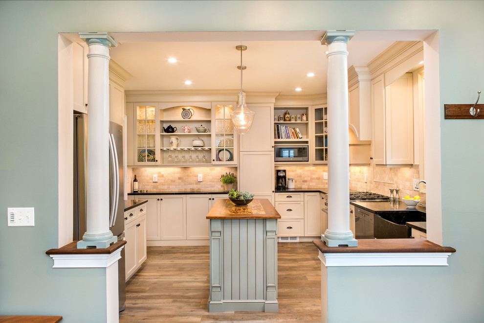 Design ideas for a mid-sized traditional separate kitchen in New York with a farmhouse sink, recessed-panel cabinets, beige cabinets, soapstone benchtops, beige splashback, travertine splashback, stainless steel appliances, vinyl floors, with island, brown floor and black benchtop.