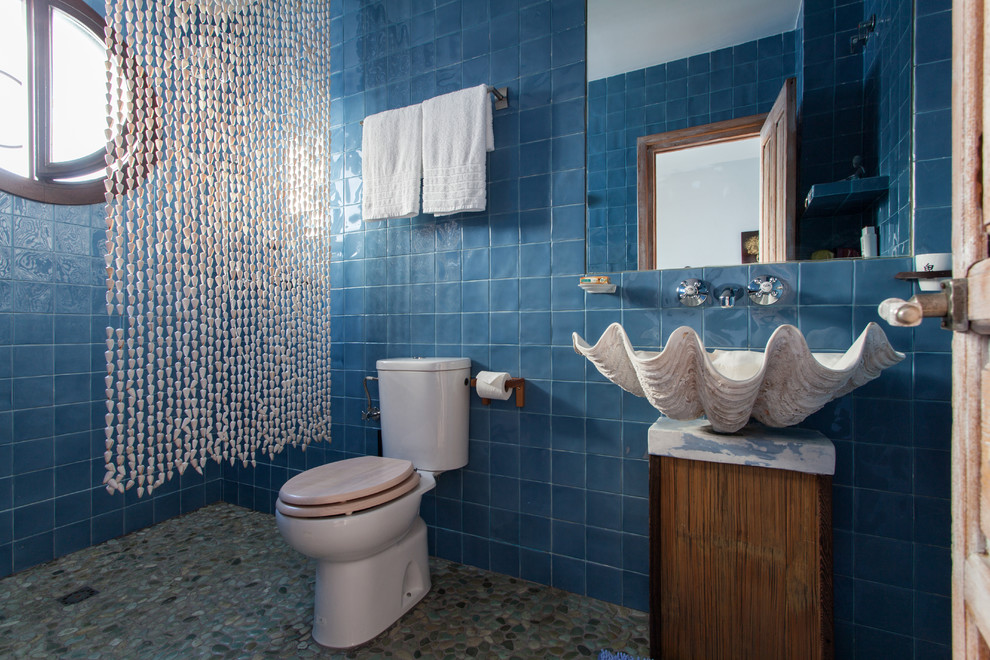 This is an example of a mid-sized tropical 3/4 bathroom in Seville with a vessel sink, a two-piece toilet, blue tile, an open shower, ceramic tile, blue walls, pebble tile floors, marble benchtops, medium wood cabinets, a shower curtain and flat-panel cabinets.