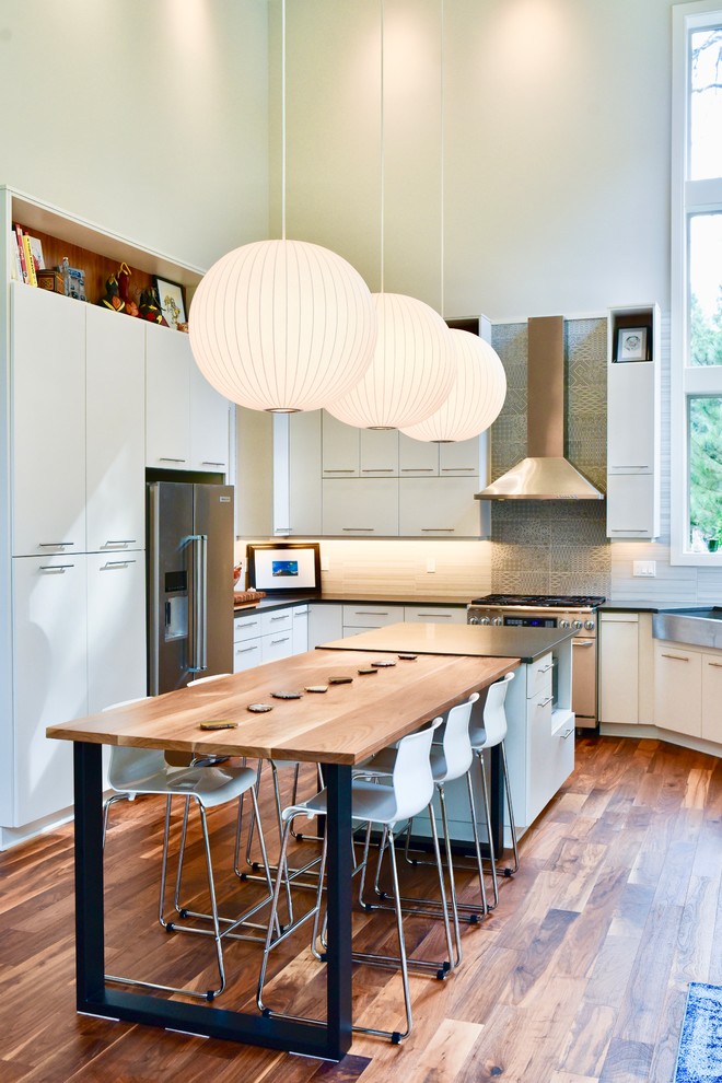 Contemporary u-shaped kitchen in Portland with a farmhouse sink, flat-panel cabinets, white cabinets, grey splashback, porcelain splashback, stainless steel appliances, medium hardwood floors, with island, grey benchtop and brown floor.
