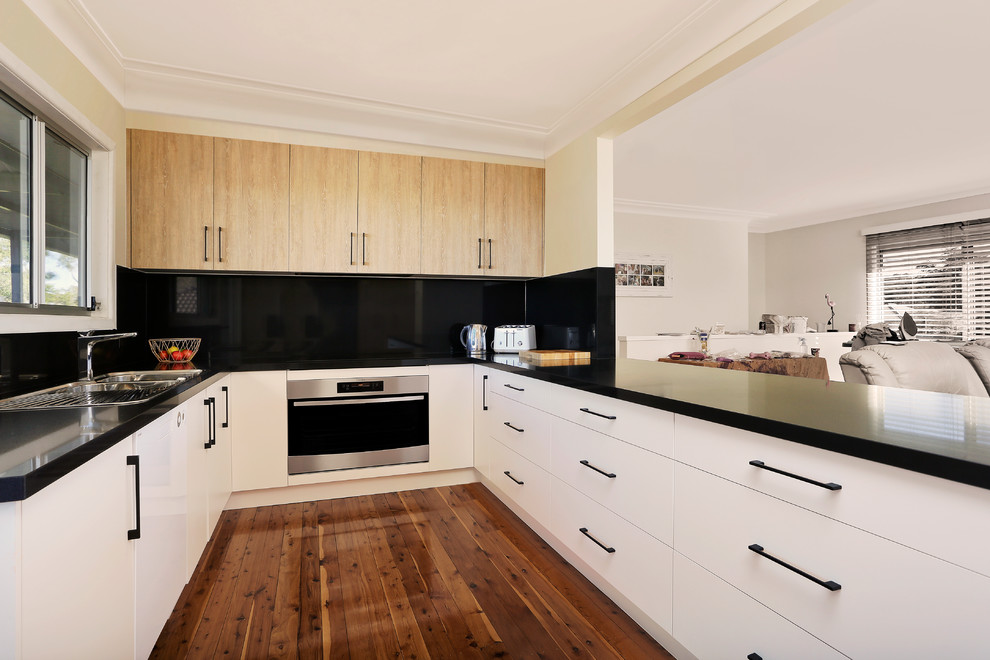 Design ideas for a mid-sized contemporary u-shaped open plan kitchen in Sydney with a drop-in sink, flat-panel cabinets, white cabinets, quartz benchtops, black splashback, stone slab splashback, black appliances, medium hardwood floors, a peninsula, brown floor and black benchtop.