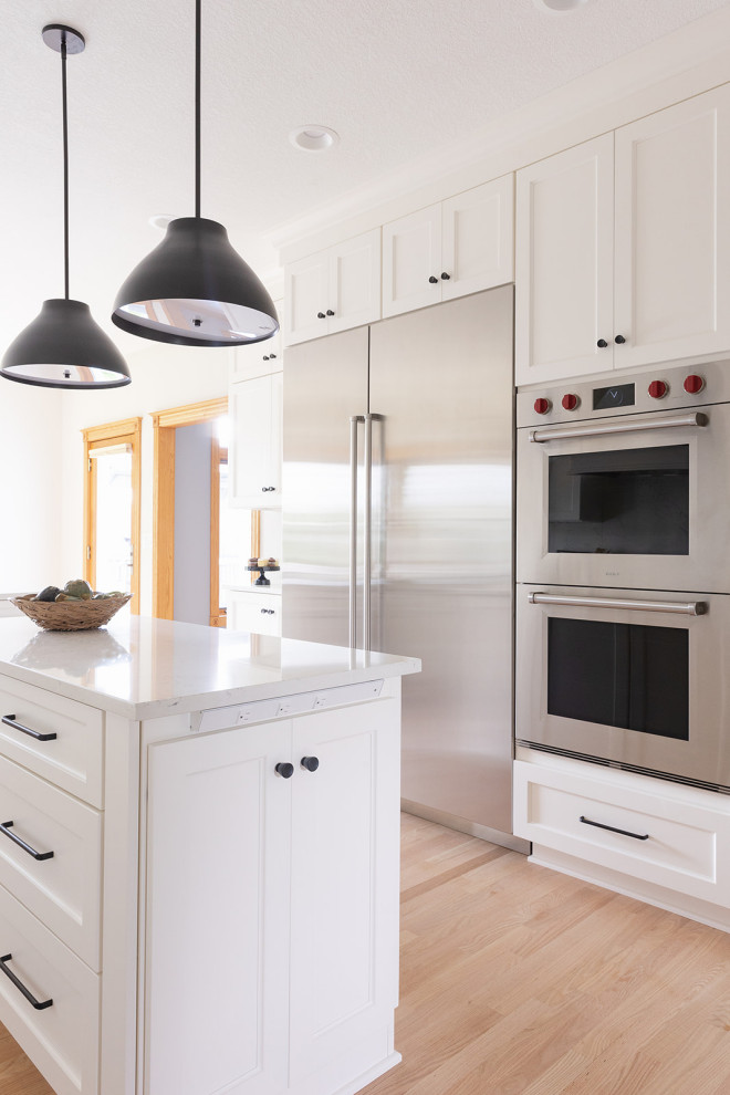 Example of a large transitional light wood floor eat-in kitchen design with an undermount sink, shaker cabinets, white cabinets, quartz countertops, white backsplash, quartz backsplash, stainless steel appliances, an island and white countertops