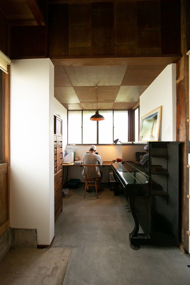 Photo of a mid-sized contemporary study room in Other with white walls, medium hardwood floors, a wood stove, a concrete fireplace surround, a built-in desk, beige floor and exposed beam.