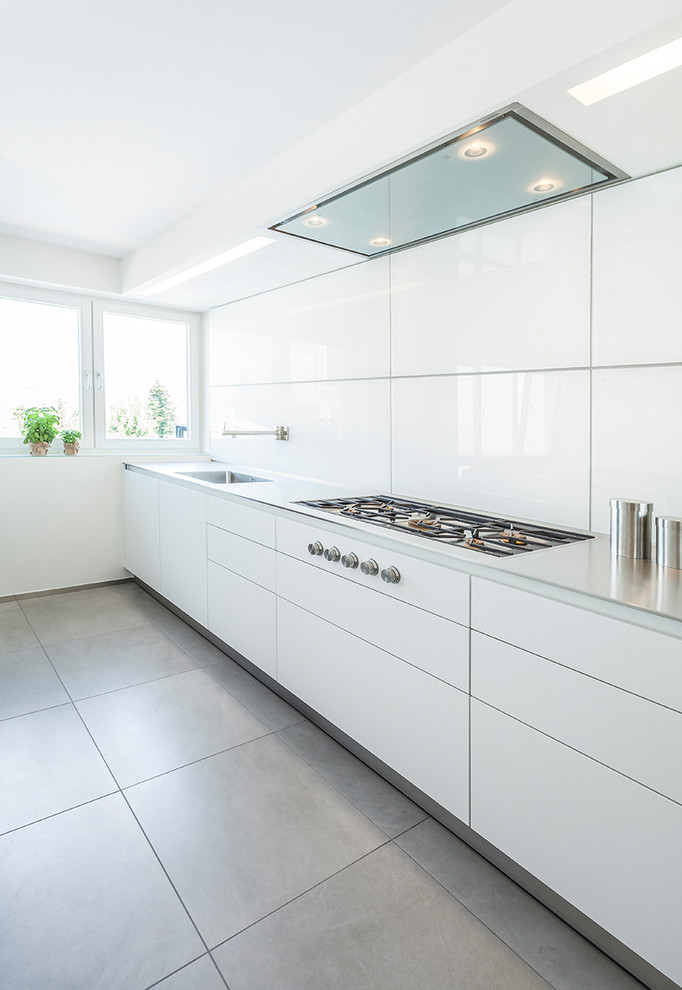 This is an example of a large contemporary single-wall kitchen in Stuttgart with a drop-in sink, flat-panel cabinets, white cabinets, stainless steel benchtops, white splashback, panelled appliances, ceramic floors and grey floor.