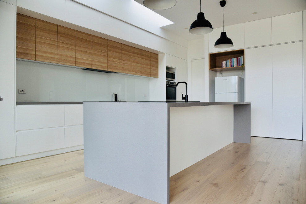 Photo of a large scandinavian galley open plan kitchen in Melbourne with a double-bowl sink, flat-panel cabinets, white cabinets, solid surface benchtops, white splashback, glass sheet splashback, stainless steel appliances, light hardwood floors, with island, beige floor, grey benchtop and recessed.