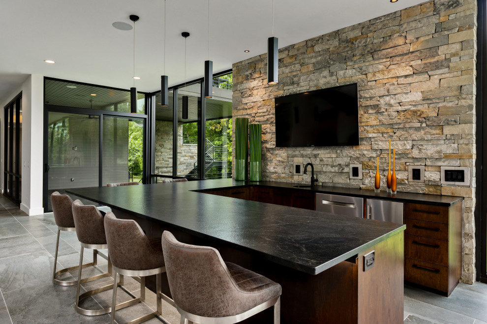 Photo of a contemporary u-shaped seated home bar in Kansas City with an undermount sink, flat-panel cabinets, medium wood cabinets, multi-coloured splashback, stone tile splashback, grey floor and black benchtop.