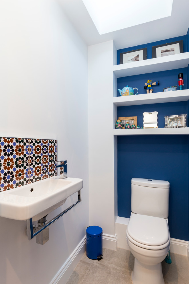 Inspiration for a small transitional powder room in London with a two-piece toilet, multi-coloured tile, blue walls, a wall-mount sink and white cabinets.