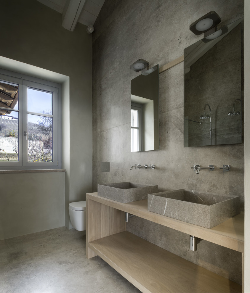 This is an example of a modern bathroom in Other with open cabinets, light wood cabinets, a wall-mount toilet, grey walls, concrete floors, a vessel sink and grey floor.