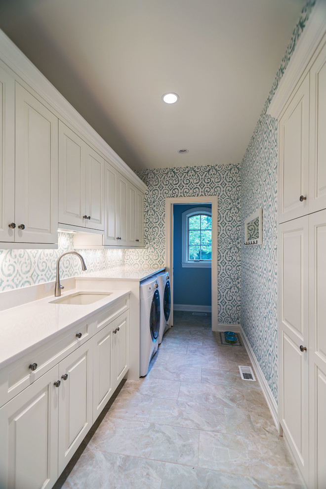 Photo of a large traditional single-wall dedicated laundry room in Cincinnati with an undermount sink, raised-panel cabinets, white cabinets, quartz benchtops, multi-coloured walls, porcelain floors, a side-by-side washer and dryer, beige floor and white benchtop.