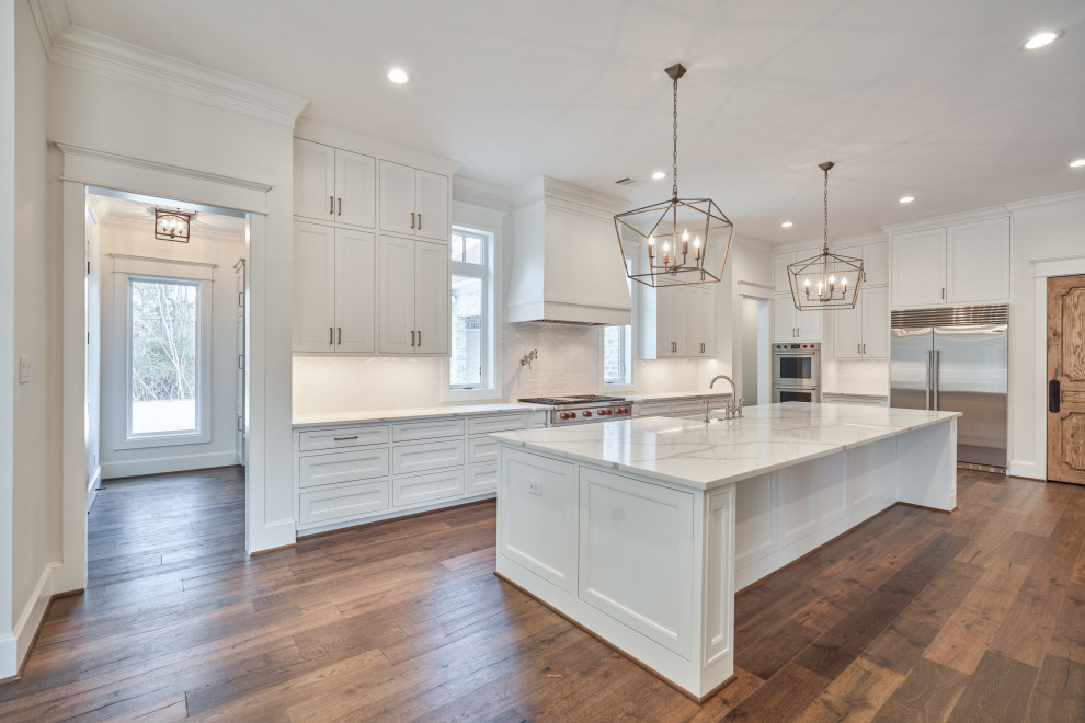 Large l-shaped open plan kitchen in Houston with recessed-panel cabinets, white cabinets, white splashback, stainless steel appliances, dark hardwood floors, with island, brown floor and white benchtop.