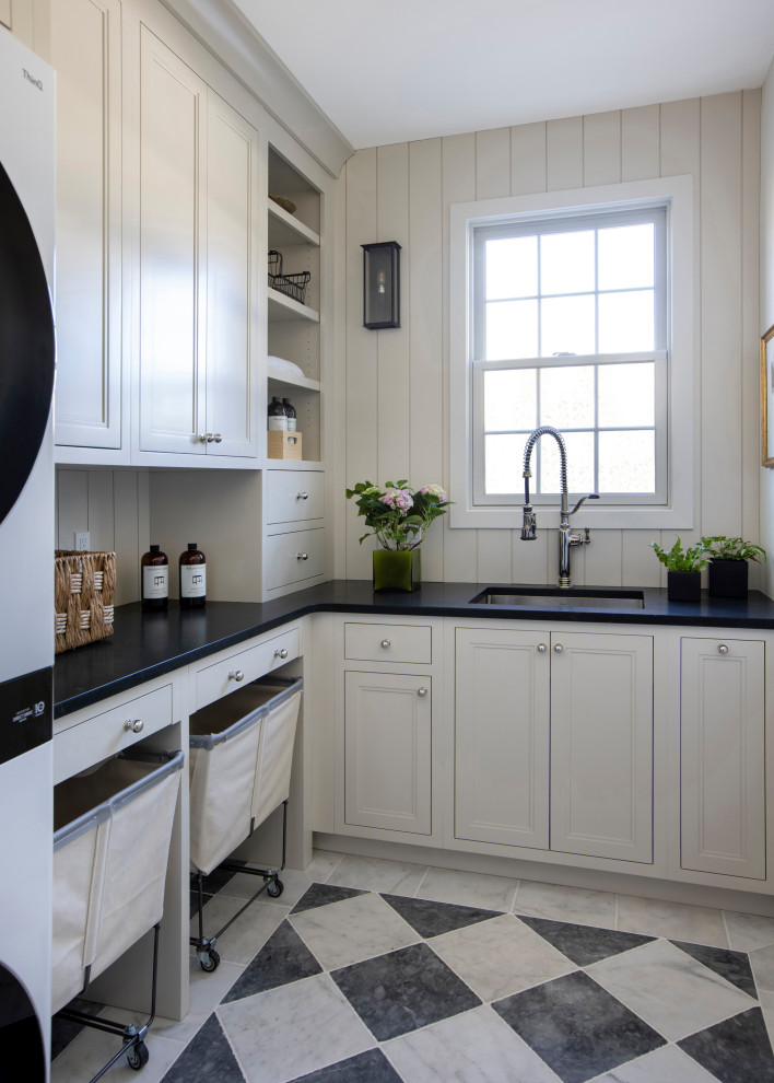 This is an example of a mid-sized transitional l-shaped dedicated laundry room in Philadelphia with an undermount sink, flat-panel cabinets, white cabinets, granite benchtops, white splashback, shiplap splashback, white walls, porcelain floors, a stacked washer and dryer, black floor, black benchtop and planked wall panelling.