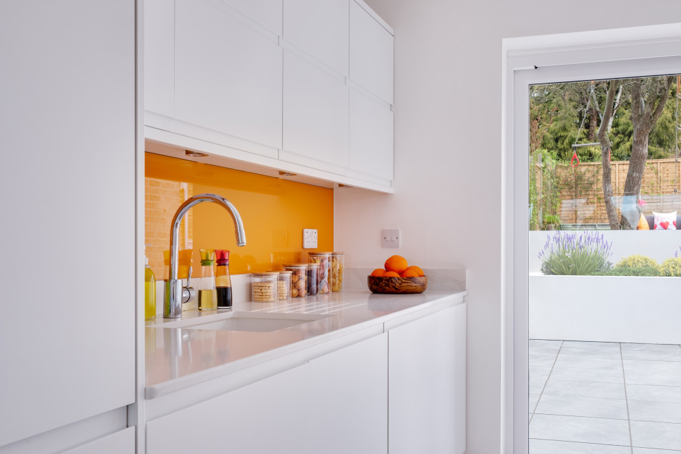 Photo of a mid-sized contemporary galley open plan kitchen in Gloucestershire with an integrated sink, white cabinets, quartz benchtops, orange splashback, glass sheet splashback, panelled appliances, porcelain floors, with island, beige floor and white benchtop.