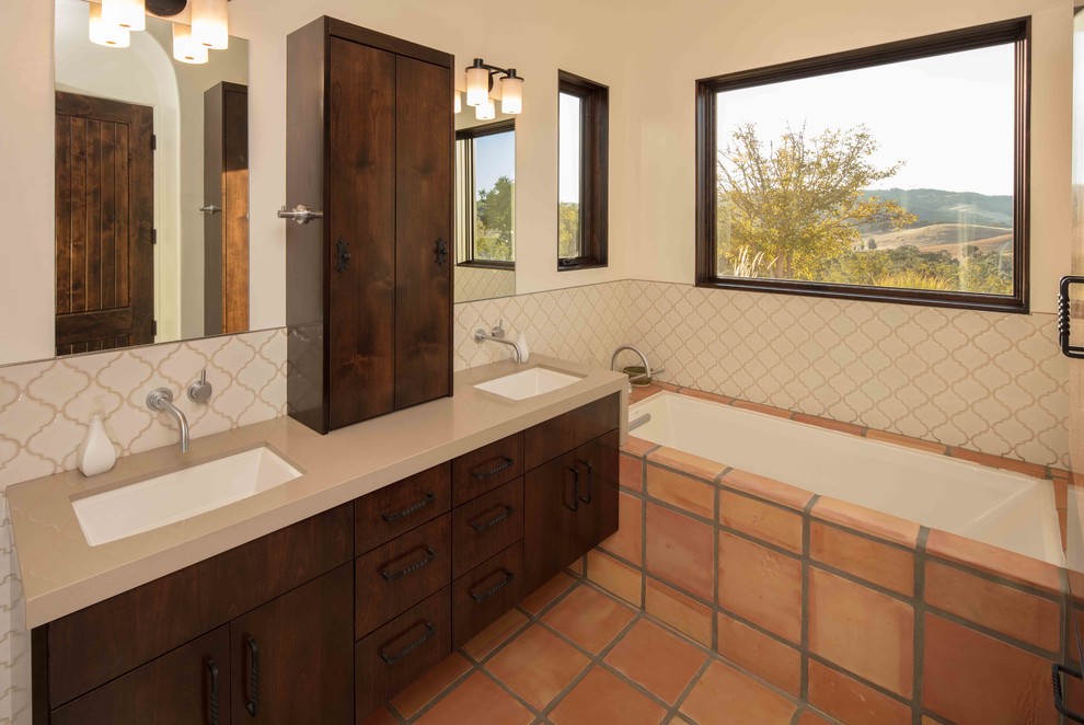 Mid-sized mediterranean bathroom in San Luis Obispo with brown cabinets, a one-piece toilet, beige tile, ceramic tile, beige walls, solid surface benchtops, a hinged shower door, flat-panel cabinets, a drop-in tub, an alcove shower, terra-cotta floors, an undermount sink and orange floor.