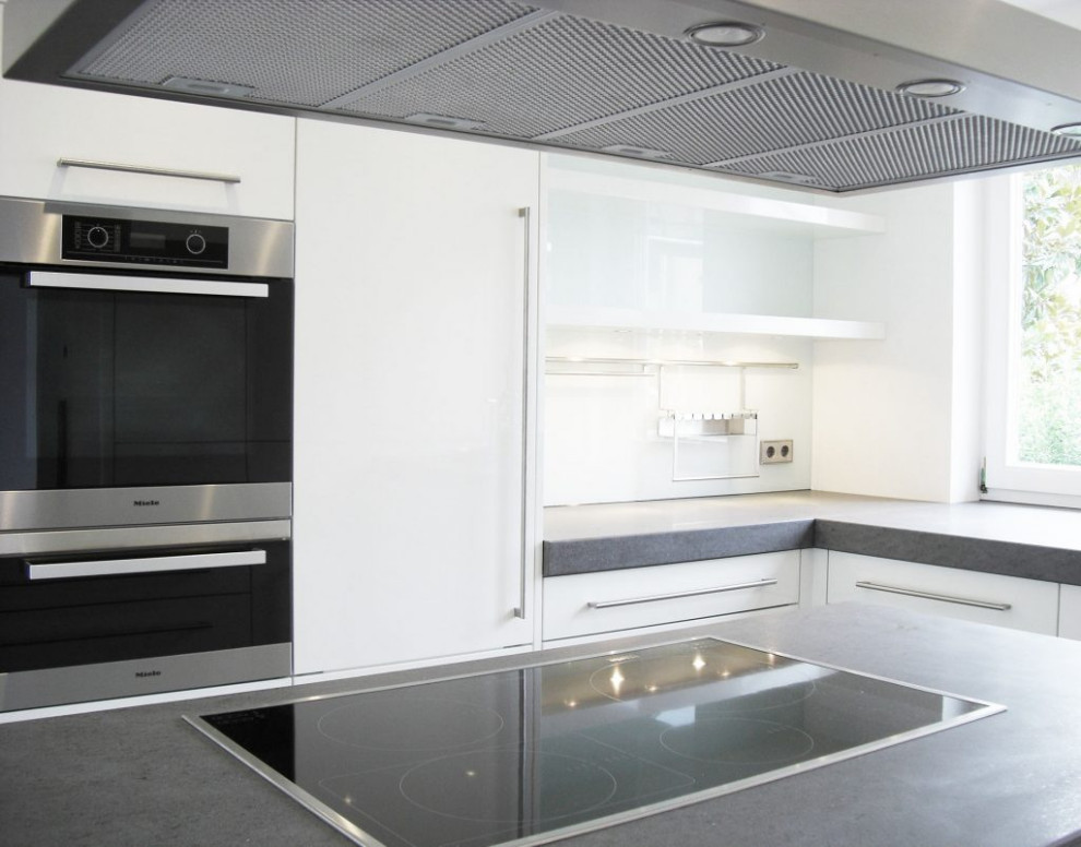 Expansive contemporary l-shaped kitchen in Other with an integrated sink, flat-panel cabinets, white cabinets, concrete benchtops, white splashback, black appliances, concrete floors, with island, grey floor and grey benchtop.
