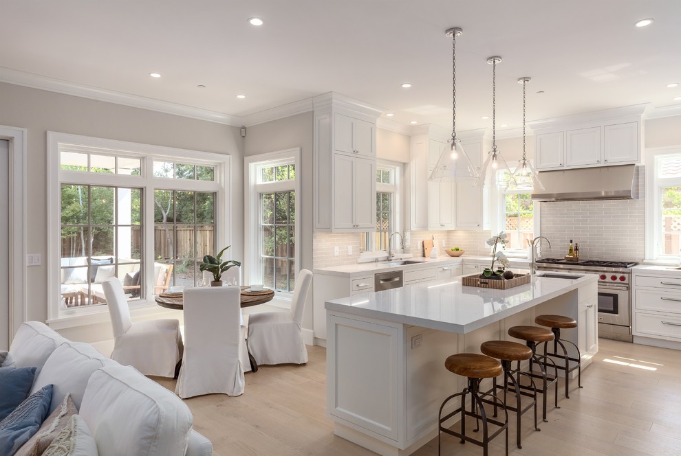 This is an example of a transitional u-shaped open plan kitchen in San Francisco with an undermount sink, recessed-panel cabinets, white cabinets, white splashback, stainless steel appliances, light hardwood floors, with island and beige floor.