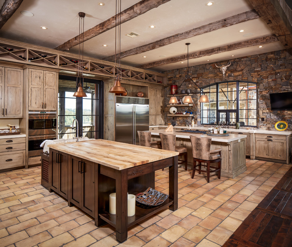 This is an example of a large country l-shaped eat-in kitchen in Houston with a farmhouse sink, raised-panel cabinets, light wood cabinets, wood benchtops, multi-coloured splashback, brick splashback, stainless steel appliances, brick floors, multiple islands and beige floor.