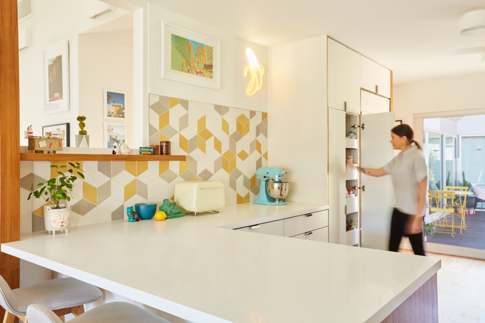 This is an example of an eclectic l-shaped eat-in kitchen in Los Angeles with a single-bowl sink, flat-panel cabinets, white cabinets, quartz benchtops, yellow splashback, ceramic splashback, stainless steel appliances, light hardwood floors, a peninsula, brown floor and white benchtop.