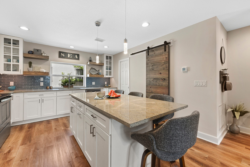 Mid-sized beach style l-shaped open plan kitchen in Jacksonville with shaker cabinets, white cabinets, blue splashback, glass tile splashback, stainless steel appliances and with island.