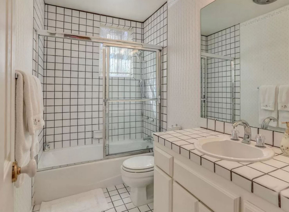 This is an example of a mid-sized country kids bathroom in San Diego with raised-panel cabinets, white cabinets, an alcove tub, a shower/bathtub combo, a two-piece toilet, white tile, ceramic tile, white walls, mosaic tile floors, an undermount sink, tile benchtops, white floor, a sliding shower screen, white benchtops, a single vanity and a built-in vanity.