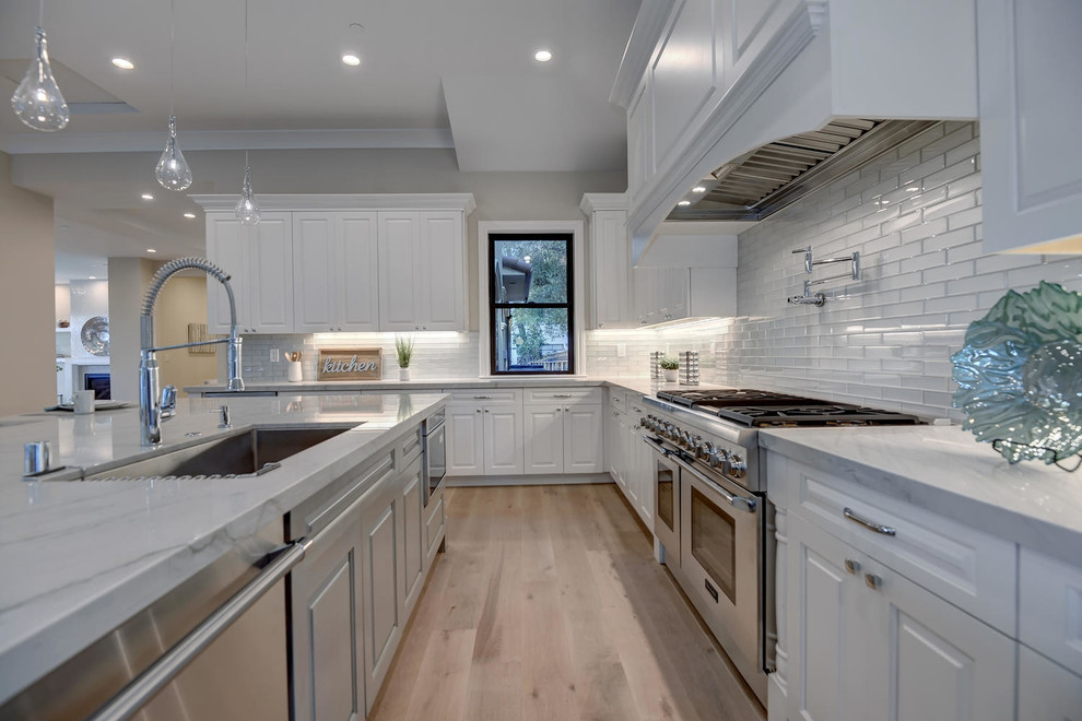 Transitional l-shaped kitchen in San Francisco with an undermount sink, raised-panel cabinets, white cabinets, glass tile splashback, stainless steel appliances, light hardwood floors, with island, beige floor, grey benchtop and metallic splashback.