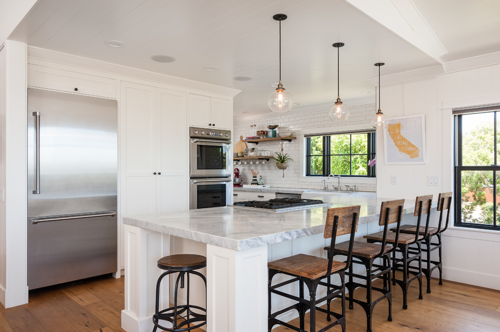 This is an example of a country kitchen in San Diego with a farmhouse sink, shaker cabinets, white cabinets, white splashback, subway tile splashback, stainless steel appliances, medium hardwood floors and a peninsula.