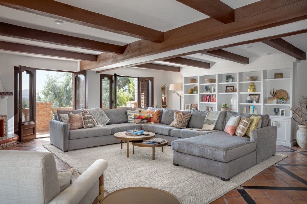 This is an example of a large mediterranean open concept family room in Orange County with a home bar, terra-cotta floors, a corner fireplace, a stone fireplace surround, a wall-mounted tv, orange floor and exposed beam.