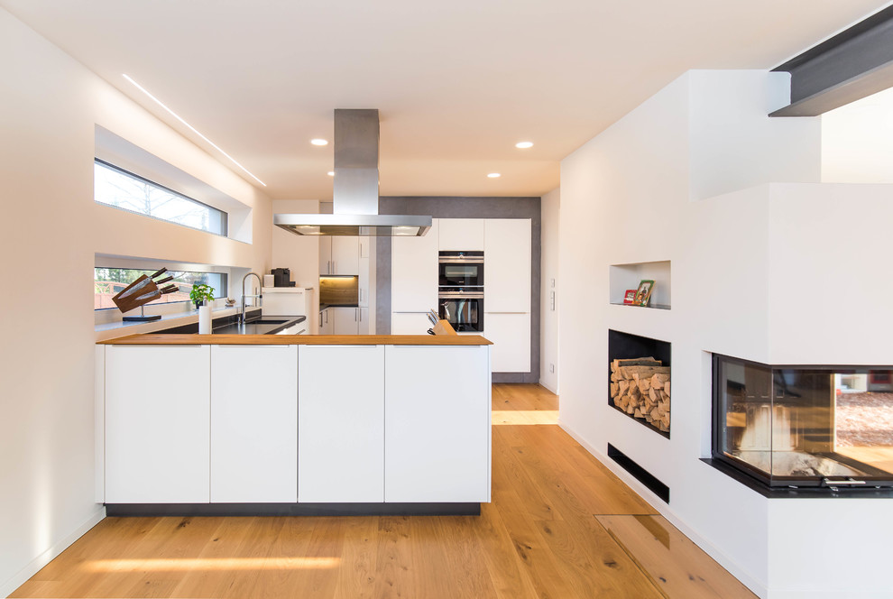 Design ideas for a contemporary u-shaped kitchen in Nuremberg with an undermount sink, flat-panel cabinets, white cabinets, wood benchtops, black appliances, medium hardwood floors, a peninsula, brown floor and brown benchtop.
