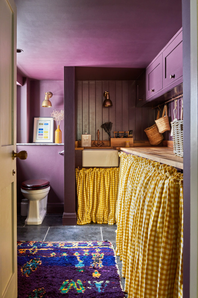 Design ideas for a mid-sized transitional l-shaped utility room in London with a farmhouse sink, shaker cabinets, yellow cabinets, wood benchtops, shiplap splashback, purple walls, limestone floors, a concealed washer and dryer, black floor, brown benchtop and planked wall panelling.