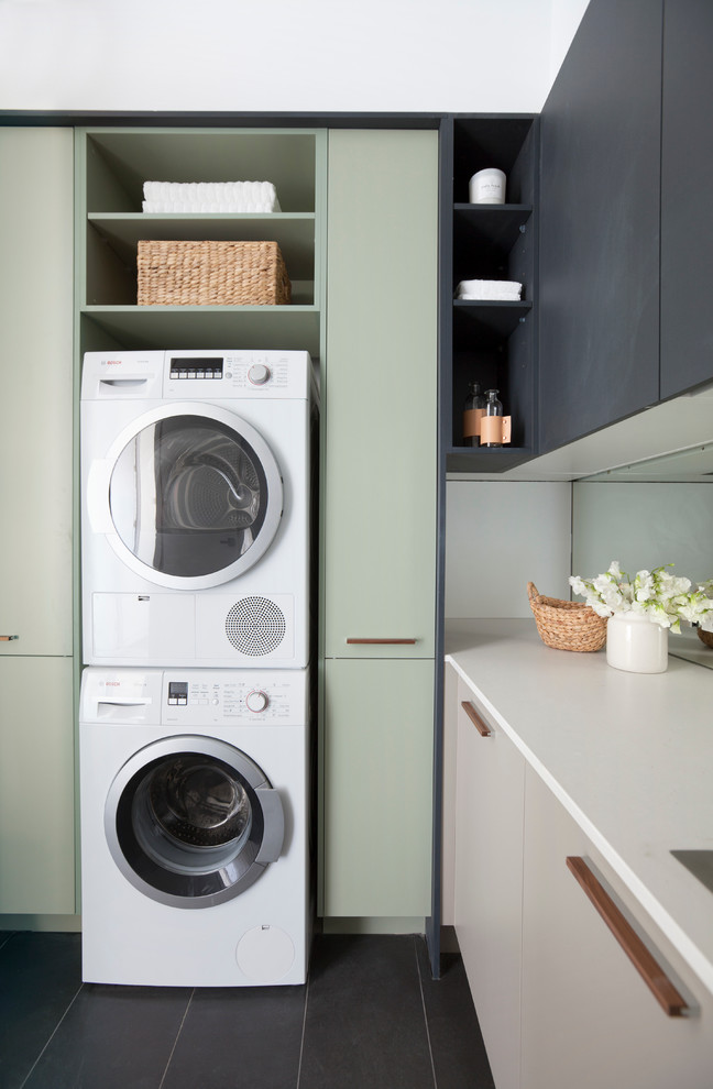 Mid-sized modern l-shaped dedicated laundry room in Melbourne with a drop-in sink, flat-panel cabinets, black cabinets, quartz benchtops, white walls and a stacked washer and dryer.