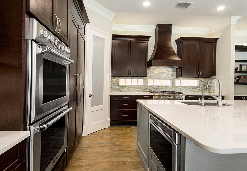 This is an example of a mid-sized transitional l-shaped open plan kitchen in DC Metro with an undermount sink, recessed-panel cabinets, dark wood cabinets, granite benchtops, beige splashback, ceramic splashback, stainless steel appliances, medium hardwood floors, with island, brown floor and beige benchtop.