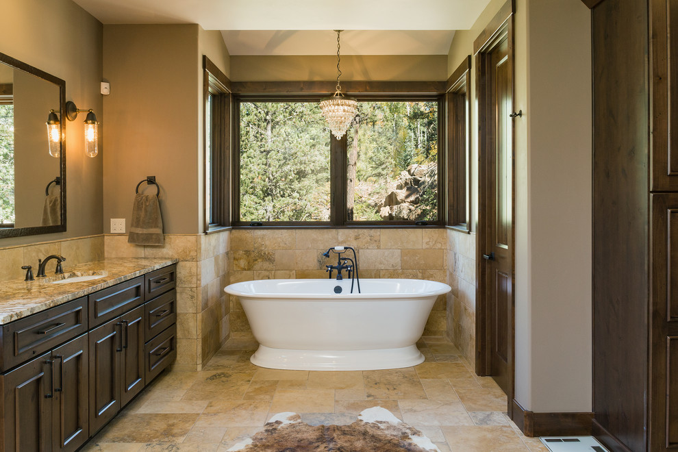 Example of a large mountain style master beige tile and stone tile travertine floor and beige floor alcove bathtub design with brown cabinets, an undermount sink, granite countertops, a hinged shower door and beige countertops