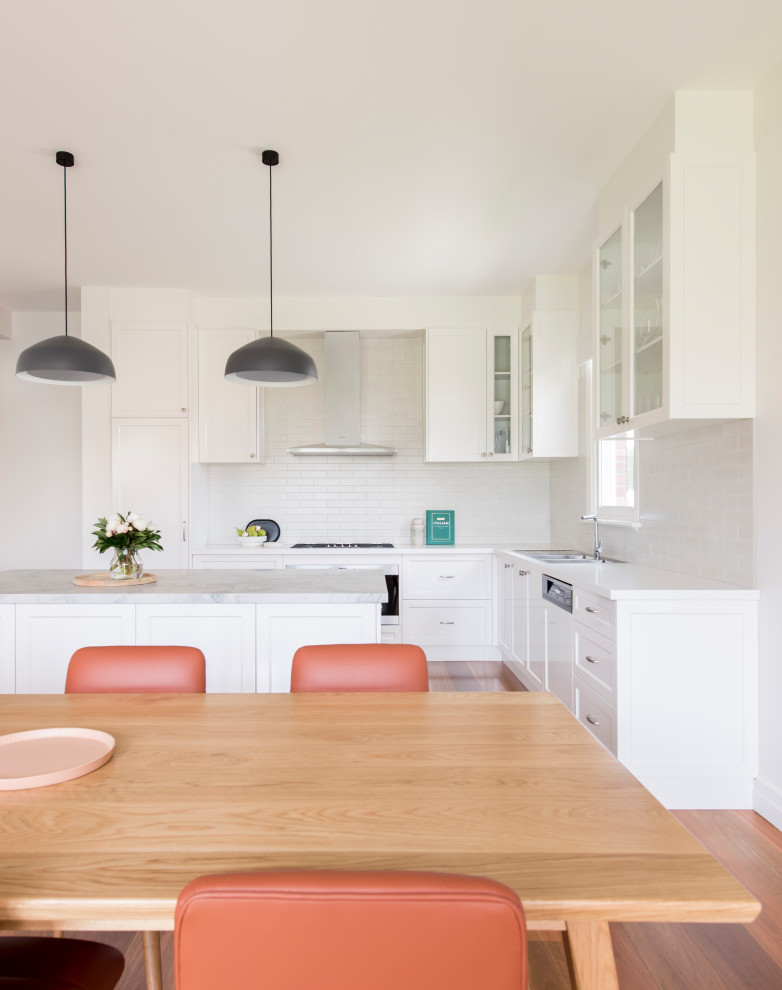 This is an example of a large contemporary single-wall eat-in kitchen in Melbourne with a drop-in sink, raised-panel cabinets, white cabinets, quartz benchtops, white splashback, subway tile splashback, black appliances, medium hardwood floors, with island, brown floor and white benchtop.