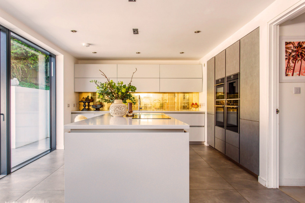Example of a mid-sized trendy l-shaped concrete floor and gray floor eat-in kitchen design in Sussex with a drop-in sink, flat-panel cabinets, white cabinets, solid surface countertops, multicolored backsplash, glass sheet backsplash, paneled appliances, an island and white countertops