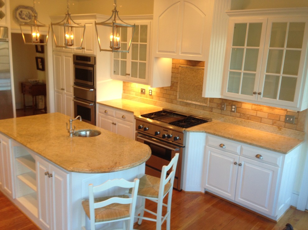 Example of a mid-sized transitional l-shaped medium tone wood floor enclosed kitchen design with an undermount sink, raised-panel cabinets, white cabinets, granite countertops, beige backsplash, stone tile backsplash, stainless steel appliances and an island