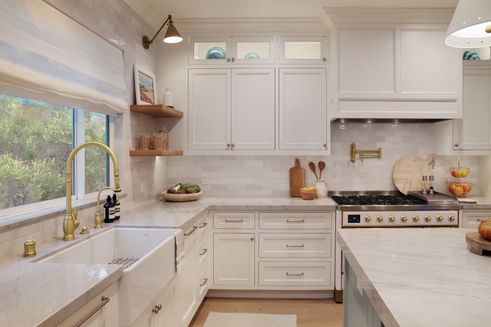 Design ideas for a large beach style l-shaped open plan kitchen in Orange County with a farmhouse sink, shaker cabinets, white cabinets, quartzite benchtops, white splashback, ceramic splashback, panelled appliances, light hardwood floors and with island.