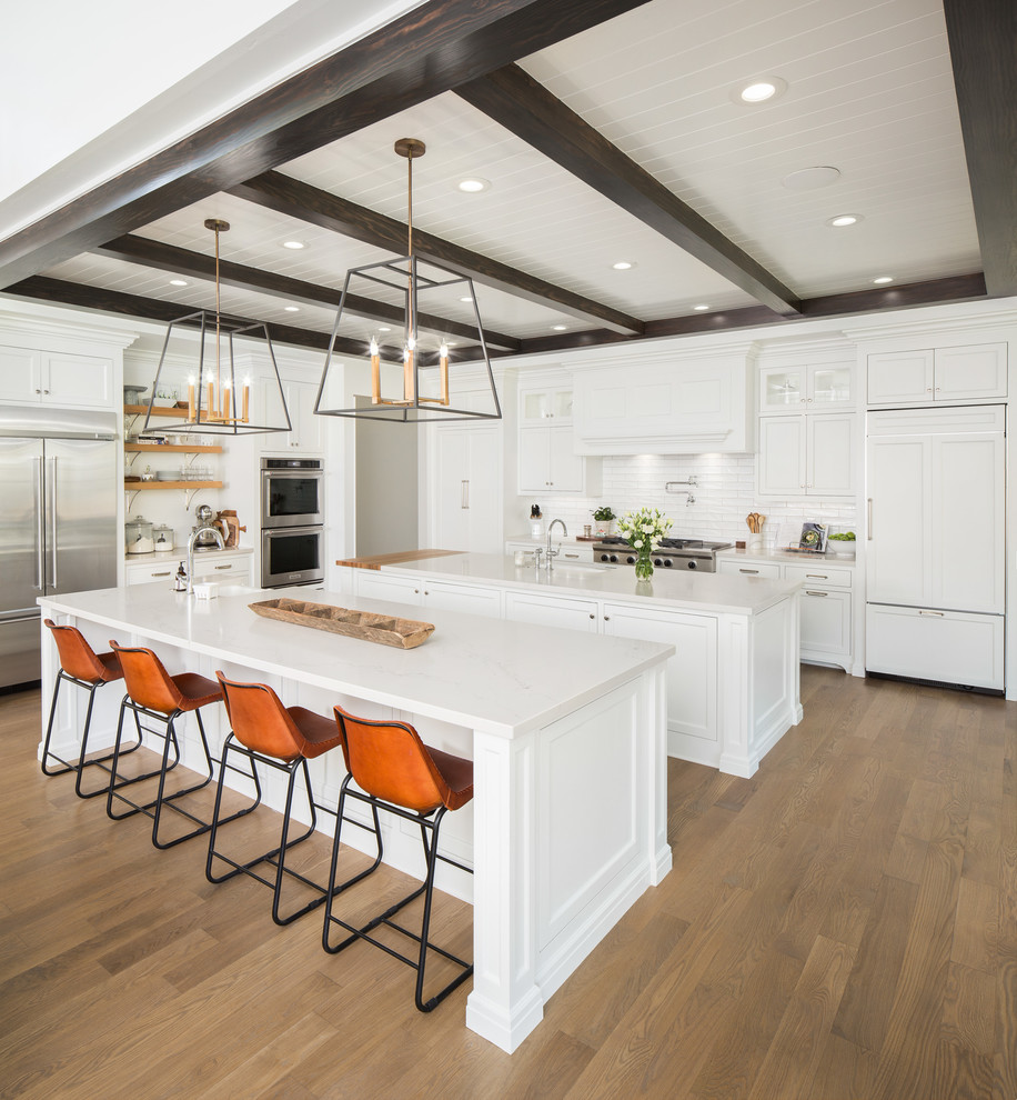 This is an example of a country l-shaped kitchen in Salt Lake City with shaker cabinets, white cabinets, white splashback, subway tile splashback, panelled appliances, medium hardwood floors, multiple islands and brown floor.