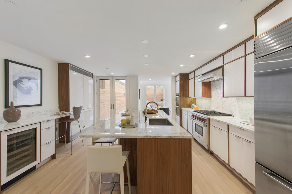 This is an example of a contemporary kitchen in New York with an undermount sink, flat-panel cabinets, white cabinets, white splashback, marble splashback, stainless steel appliances, light hardwood floors, with island and beige floor.