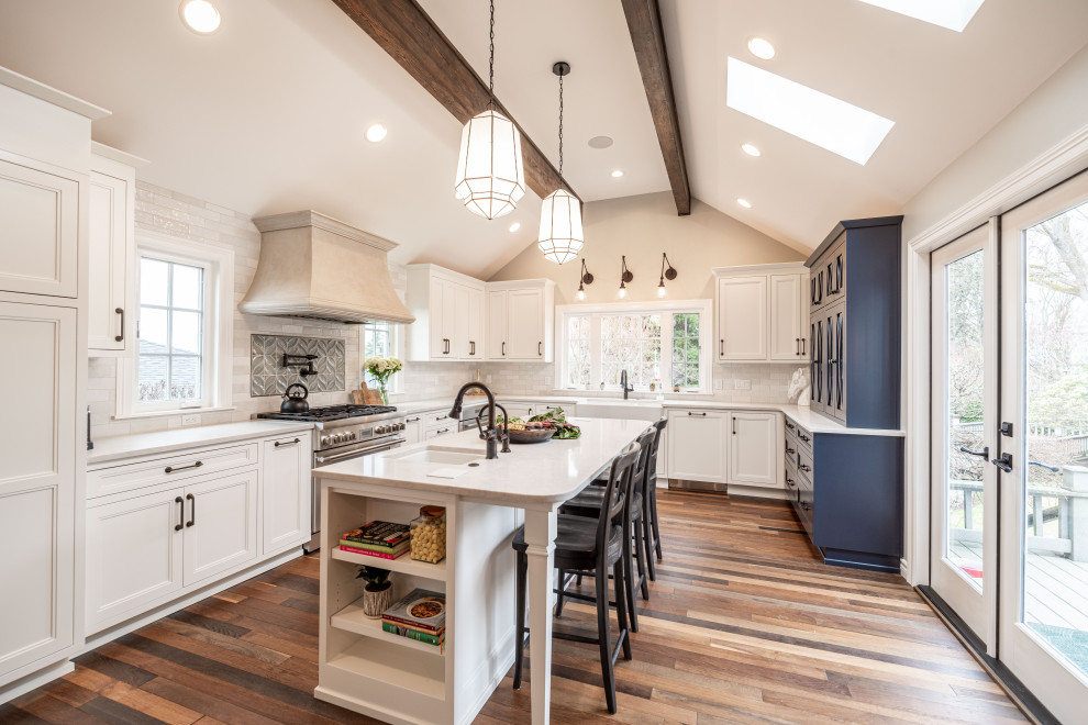 This is an example of a transitional u-shaped kitchen in Seattle with a farmhouse sink, recessed-panel cabinets, white cabinets, quartz benchtops, multi-coloured splashback, subway tile splashback, panelled appliances, medium hardwood floors, with island, brown floor, beige benchtop, exposed beam and vaulted.