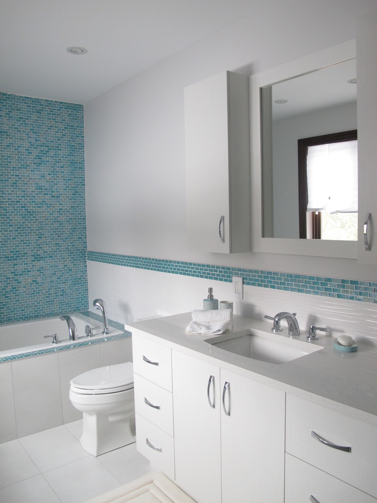 Photo of a mid-sized contemporary kids bathroom in Montreal with an undermount sink, flat-panel cabinets, white cabinets, solid surface benchtops, a drop-in tub, an alcove shower, a one-piece toilet, blue tile, mosaic tile, white walls and porcelain floors.